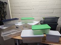 Photo showing assorted plastic storage containers including clear, white, green, and black varieties stacked on a table.