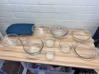 Wide shot showing all glass kitchen dishes arranged on table including bowls, baking dishes, measuring cup, and storage container with lid.