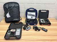 Full view of lot showing multiple electrotherapy devices, their cases, accessory cables, charger, electrodes, and carrying bag placed on wooden surface against white block wall.