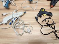 Top-down shot showing six electric hot glue guns of different colors and designs scattered on wooden surface with cords.