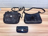 Three items on wooden surface: two black leather purses and one matching wallet, all featuring decorative stitching and silver hardware