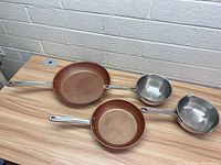 Two copper-colored non-stick frying pans and one stainless steel saucepan shown in top view on wooden table.