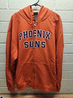 Front view of orange zip-up hoodie hanging on a wall. Shows embroidered PHOENIX SUNS lettering on chest, full zipper, hood, and front pocket.