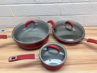 Three Pioneer Woman cookware items shown with their lids: two frying pans and one saucepan, all in red with silver interiors.