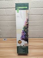 Photo of the 32-inch fiber optic tree in its original box resting on a wooden table against a white brick wall, showing the packaging label and tree image.