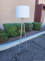 Tripod floor lamp shown outdoors with a white drum shade on top and metallic tripod legs with black feet caps, photographed from front and side angles.