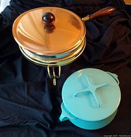 Copper fondue pot with lid and wooden handle, brass burner stand, and blue enamel pot with lid set on black cloth background.