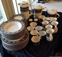 Photo shows stacked cream-colored plates with a blue floral and geometric border pattern, assorted matching mugs, and covered pots with round lids, including a coffee pot and teapot.