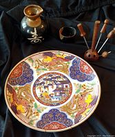 Large Asian-themed round porcelain plate with multicolor traditional designs, small black ceramic pot with calligraphy, and wooden-handled serving forks in a wooden holder on a black cloth.