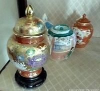 Photo showing three small Chinese vases in a row on a patterned surface, including a covered ornate vase on a wooden stand and two smaller decorative vases.