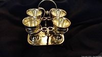 Set of 4 silver goblets in their silver holder stand on a dark cloth background, showing front and side views of the goblets and holder.