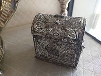 Photo showing side front view of a small silver lace box with hinged lid on a table near window light. The intricate filigree lace metal design and clasp are visible.