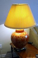 Photo of table lamp with ceramic orange base and pleated cream shade, lamp turned on showing warm light.