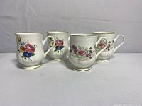 Four white ceramic cups on pedestal bases with floral patterns and gold trim shown in a group.