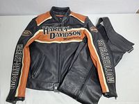 Black and brown Harley Davidson leather motorcycle jacket and black leather pants laid flat showing front and sleeve Harley Davidson logos.