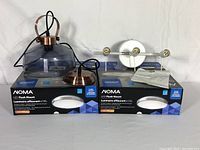 Two boxed new NOMA LED flush mount light fixtures and one hanging light fixture with smoked glass dome and copper metal accents all displayed on white background.