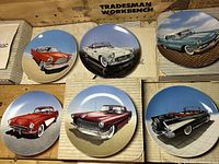 Six ceramic collector plates showing classic cars in different scenic settings, placed on a wooden surface, some in boxes.