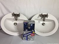 Two white oval sinks with installed Danze faucets placed side by side with unopened Moen faucet box between them.