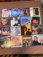 Photo showing 16 45 RPM vinyl records with colorful original sleeves laid out on a table, featuring artists Rod Stewart, Tina Turner, Kenny Rogers, and others.