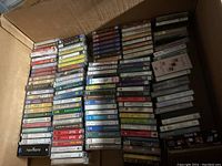 Stacked arrangement of over 120 cassette tapes in a cardboard box showing colorful cassette labels and spines