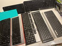 Top-down photo of seven assorted computer keyboards arranged on a beige surface. Mix includes wired and wireless types in various colors including black, gray, teal with different key styles.