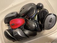 Photo showing a plastic container holding a mixed assortment of roughly a dozen computer mice in various styles, colors like black, grey, red, and silver, some branded Logitech and Microsoft.