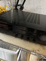 Top and front view showing ventilation slots and several control buttons on a black metal stereo receiver.