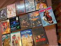 Photo shows 15 DVD movie cases arranged on a wooden floor. Titles include classics like "Pulp Fiction", "The Spy Who Loved Me", "Avatar" (multiple discs), "Sex and the City", and TV series collections like "Two and a Half Men", "Big Bang Theory", and "Eureka". Some DVDs are still sealed in plastic.