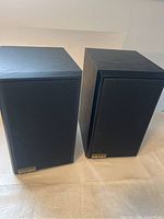 Full view of both Incus speakers with grille covers on, showing rectangular shape and black wood finish.