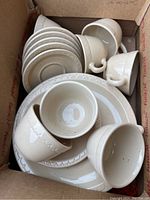 Shoebox containing stacked Syracuse Restaurant Ware, including cups, plates, saucers, and bowls in off-white color with raised border design.