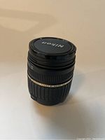 Side view of Tamron 18-200mm lens with Nikon lens cap on, showing zoom and focus grip texture and gold accent ring