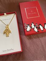 Gold tone metal necklace with geometric penguin pendant presented inside Vera Bradley branded red box featuring penguin graphics, showing necklace pendant and chain.