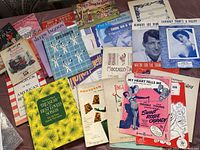 Photo of approximately 40 pieces of vintage sheet music laid out, showing variety of titles and colorful covers with different graphics and fonts.