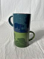 Two stacked mugs showing bear silhouettes in nature scenes, one green and one dark blue.
