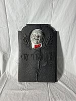 Full view of the black blowmold tombstone with raised creepy face wearing a red bow tie and text 'CRYPTIC KOOK'. Shows cracks.