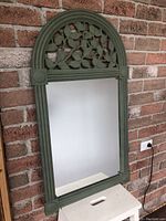 Full view of the vintage green arched mirror standing against a brick wall, showing the mirror glass and frame details.