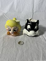 Front view of both Sammy Sam and Sylvester head mugs with a coin for scale