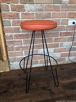 Side view of the vintage orange cushioned stool with black iron legs against a brick wall and wooden floor.