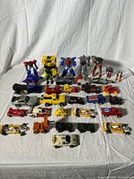 Full view of the vintage Transformers action figures and toy cars laid out on a white cloth background showing multiple items including notable robot figures and a variety of toy cars.