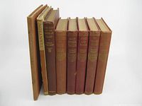 Front view of eight vintage old books standing upright showing cloth covers in reddish-brown and tan colors with gold embossed text on spines.