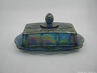 Top-front angle showing rectangular iridescent carnival glass butter dish with grapevine leaf and grape cluster motifs on lid and base. Bead textured grape cluster knob on lid. Shows wear on glass surface.