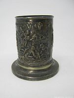 Front view of the cylindrical silverplate holder showing embossed dancing and dining scenes on a circular base with aged patina and wear marks.