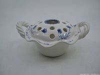 White ceramic flower frog with hand painted blue floral decorations, multiple holes on top, and twisted rope-style handles on each side.