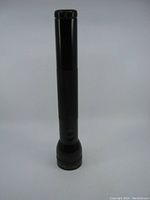 Front view of vintage black Mag-Lite flashlight standing upright on a white background, showing full length and details of the flashlight body and head.