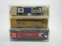 Three boxed/vintage model trains arranged stacked: bottom red Bachmann CP Rail engine in box, middle yellow Burpoh Beer rail car, top blue 1993 Holiday Box Car.