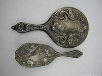 Top view of hand mirror and brush showing ornate floral and scroll silver plate designs on backs.