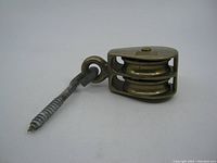 Side view of old solid brass double sheave fixed pulley showing brass sheaves and metal screw hook attached.