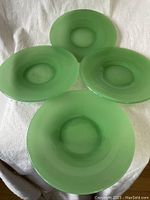 Four vintage green jadeite bread plates laid flat on white fabric, showing top views with circular center depression.
