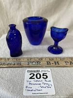 Three cobalt blue glass items: a fish shaped jar, a rounded bowl, and a goblet style candle holder with a measuring ruler and lot tag.