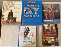 Top view showing the five books arranged on a cloth, including 'Home Body' by Joanna Gaines, 'Complete DIY Manual' by Collins, 'Measure Twice' by Bryan Baeumler, 'Lovable Livable Home' by Sherry & John Petersik, and 'The Upholsterer's Step-by-Step Handbook' by Alex Law.